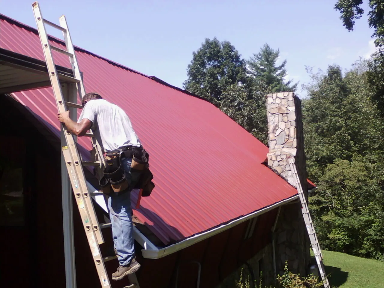 Roofer on a ladder beside a red metal roof and stone chimney — Soffit & Fascia Repair by Roofing Brothers in Bellmawr
